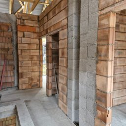 Paweł Wąchała PW usługi remontowo-budowlane - Wnętrze budynku w trakcie budowy: ściany z bloczków ceramicznych i betonowych, widoczne stropy drewniane. W tle postać w drzwiach. Surowy stan, brak wykończenia.