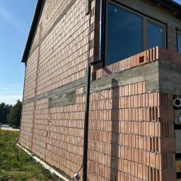 Paweł Wąchała PW usługi remontowo-budowlane - Wznoszenie ściany z czerwonej cegły dziurawki na budowie domu jednorodzinnego, widoczne okna i fragment dachu. Na pierwszym planie trawa i studzienka kanalizacyjna.