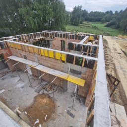 Paweł Wąchała PW usługi remontowo-budowlane - Budowa domu z cegły: zbrojenie wieńca i ściany, żółte szalunki. Widok z góry na plac budowy z zielonym tłem.