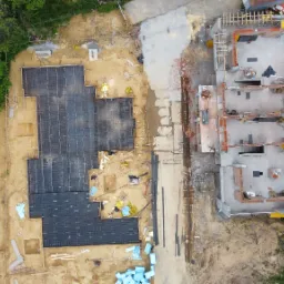 Widok z drona na plac budowy: fundamenty zbrojone na czarno i mury parteru domu z cegły, otoczone zielenią i piaskiem, ukazujące etap prac budowlanych.