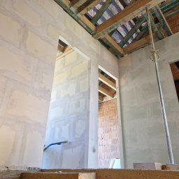 Paweł Wąchała PW usługi remontowo-budowlane - Wnętrze domu w trakcie budowy: surowe ściany z widocznymi warstwami tynku, drewniana konstrukcja dachu i ceglane ściany w tle. Wspornik podtrzymujący strop.