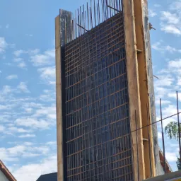 Szczeg&oacute;łowe ujęcie zbrojenia ściany w drewnianym szalunku, przygotowanego do zalania betonem na tle błękitnego nieba z obłokami.