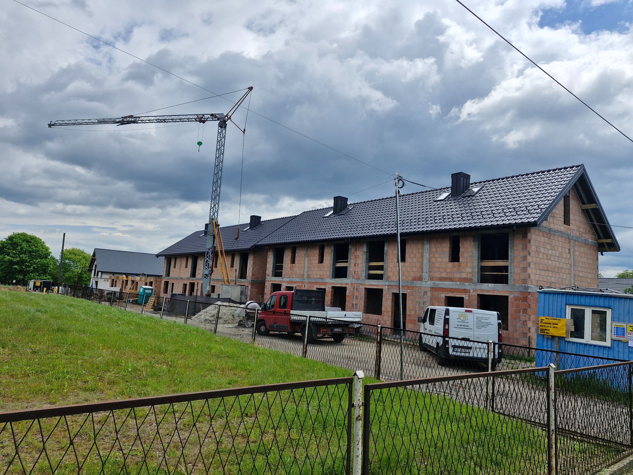 Szeregówka w stanie surowym z cegły, z dachem z czarnej dachówki, widoczny żuraw budowlany i samochody dostawcze. Ujęcie zza metalowego ogrodzenia.