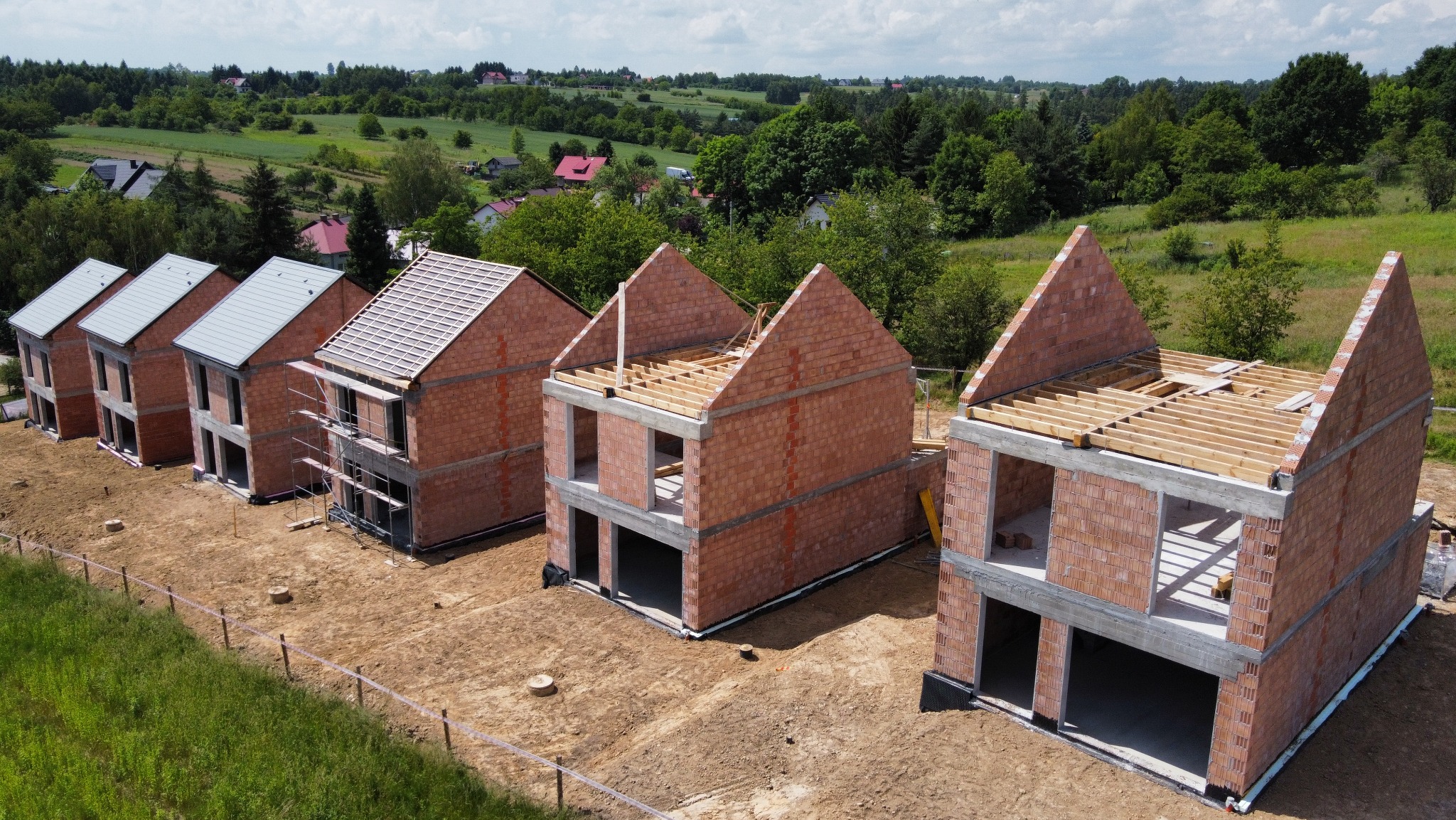 Rząd domów w trakcie budowy, z różnym stopniem ukończenia: od fundamentów i ścian po dach. Widok z góry, otoczenie zielone, częściowo zalesione.