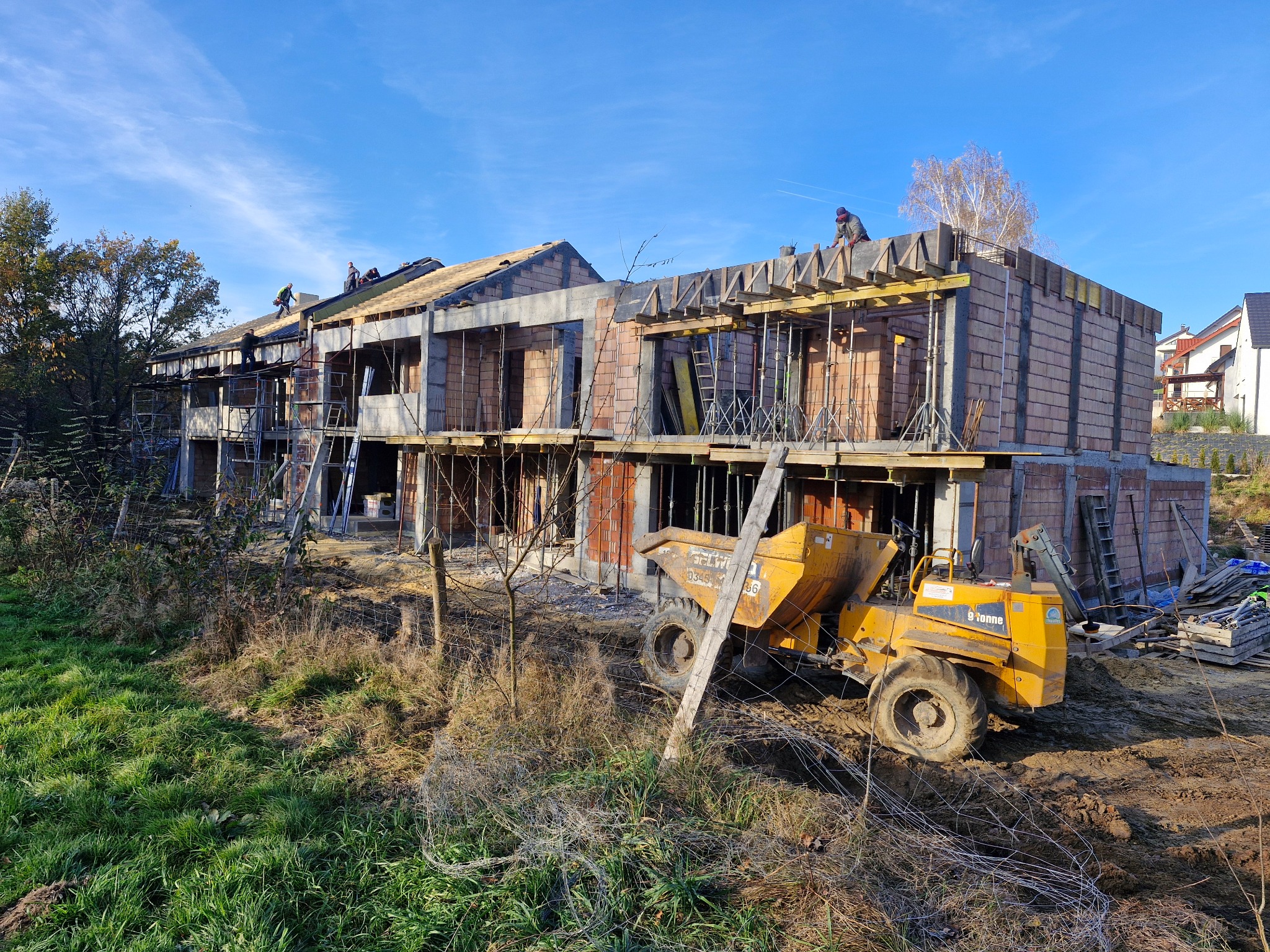 Dwukondygnacyjny dom w trakcie budowy z betonu komórkowego, z widocznym żółtym wywrotnym wozidłem i pracującymi na dachu robotnikami w słoneczny dzień.