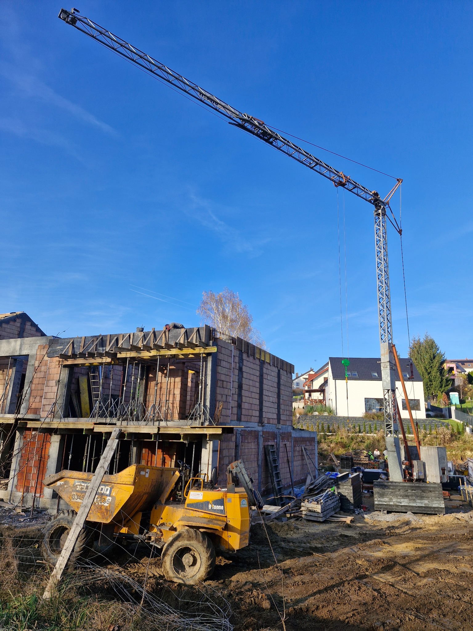 Budowa domu w stanie surowym otwartym, z widocznym żurawiem i wywrotką na tle błękitnego nieba. W tle zabudowa jednorodzinna i drzewa.
