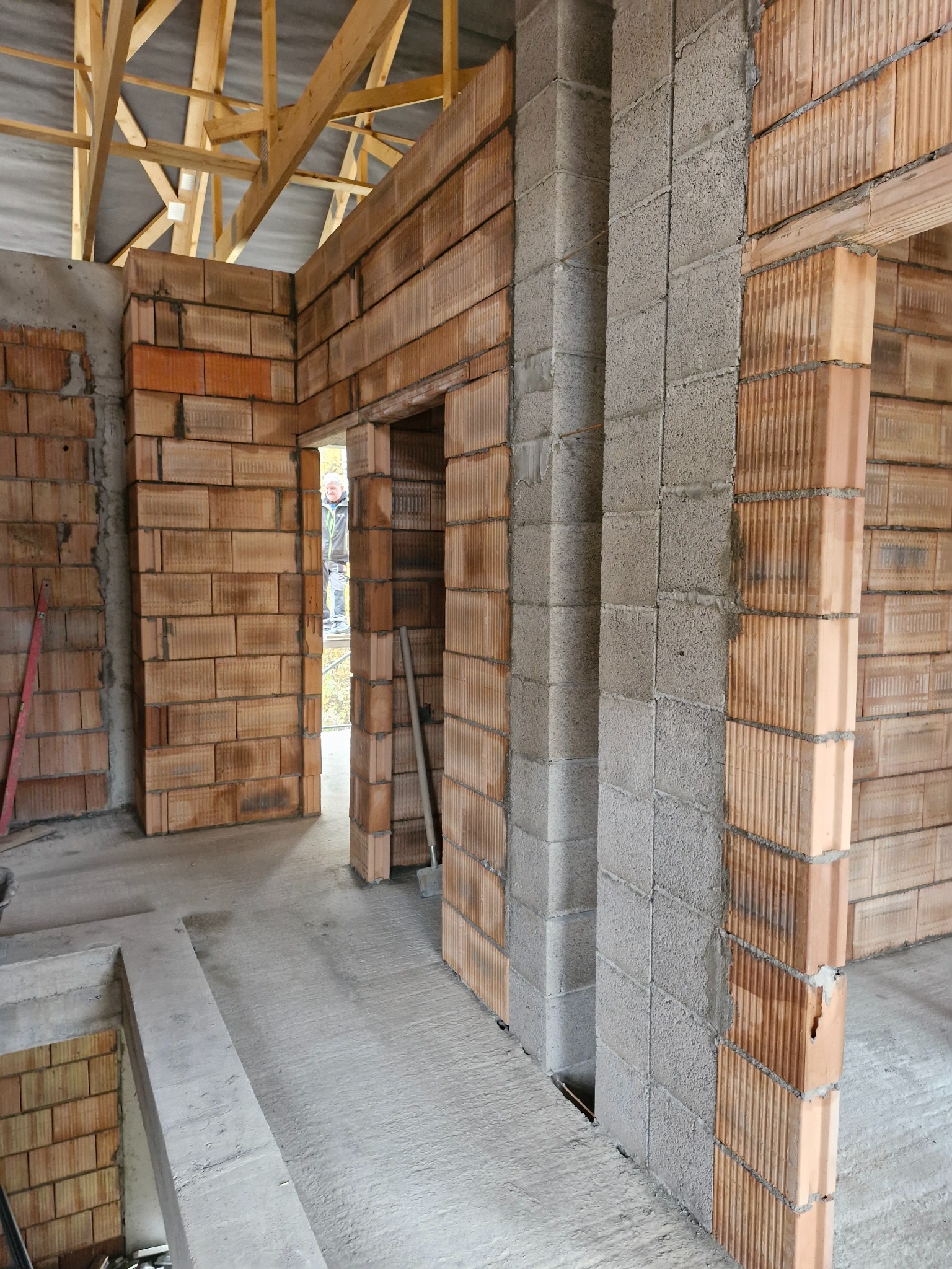 Wnętrze budynku w trakcie budowy: ściany z bloczków ceramicznych i betonowych, widoczne stropy drewniane. W tle postać w drzwiach. Surowy stan, brak wykończenia.