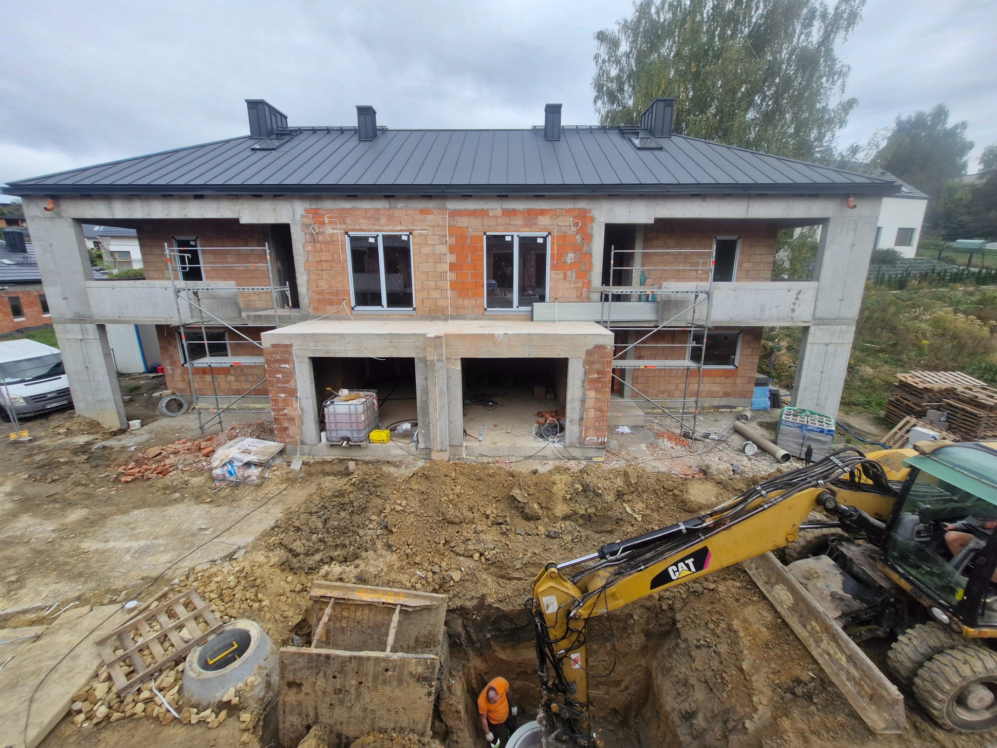 Budowa bliźniaka: surowy stan z oknami, rusztowania, wykop z koparką CAT i pracownikiem w pomarańczowej koszulce. Widok z góry na plac budowy.