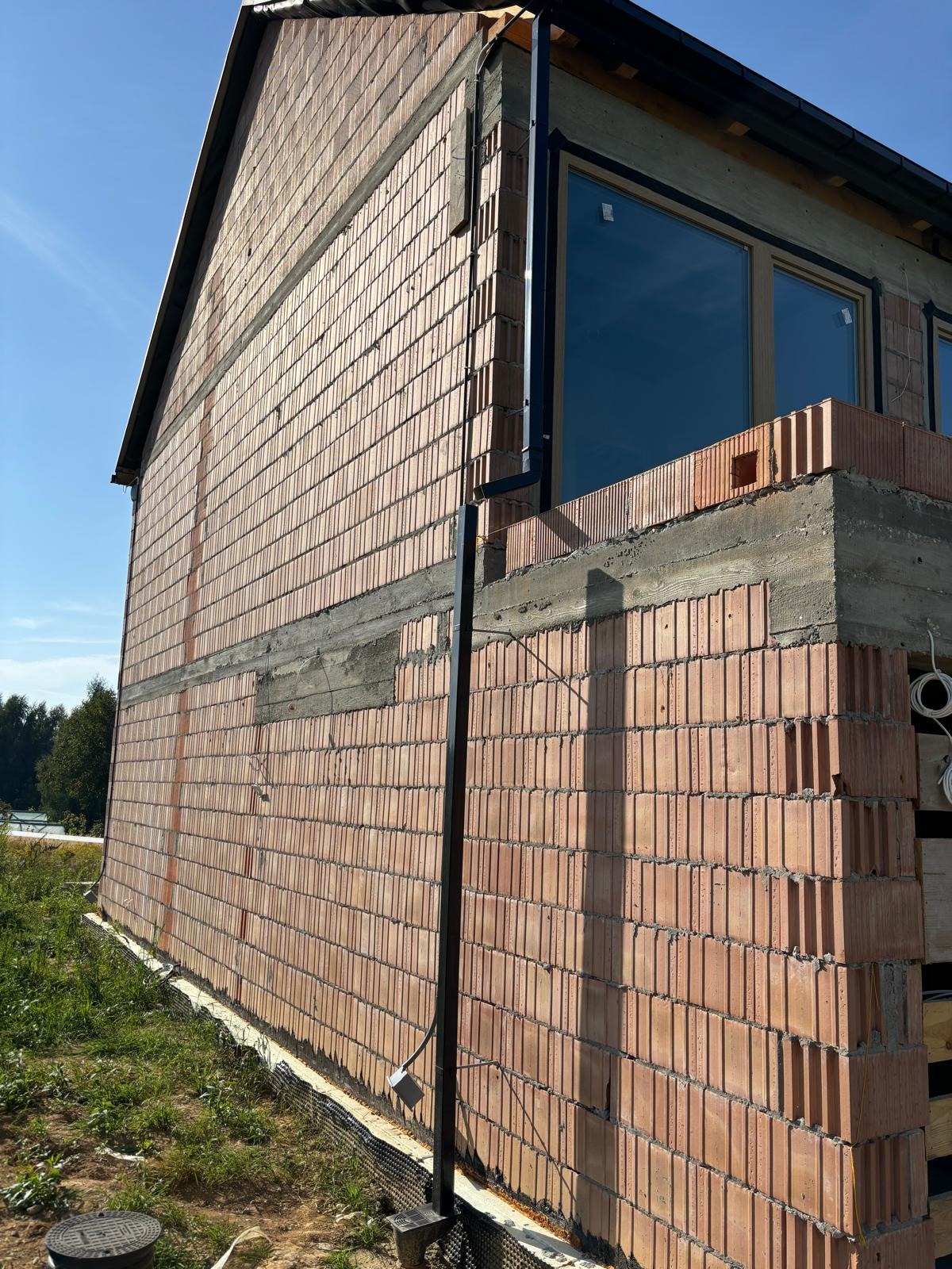 Wznoszenie ściany z czerwonej cegły dziurawki na budowie domu jednorodzinnego, widoczne okna i fragment dachu. Na pierwszym planie trawa i studzienka kanalizacyjna.