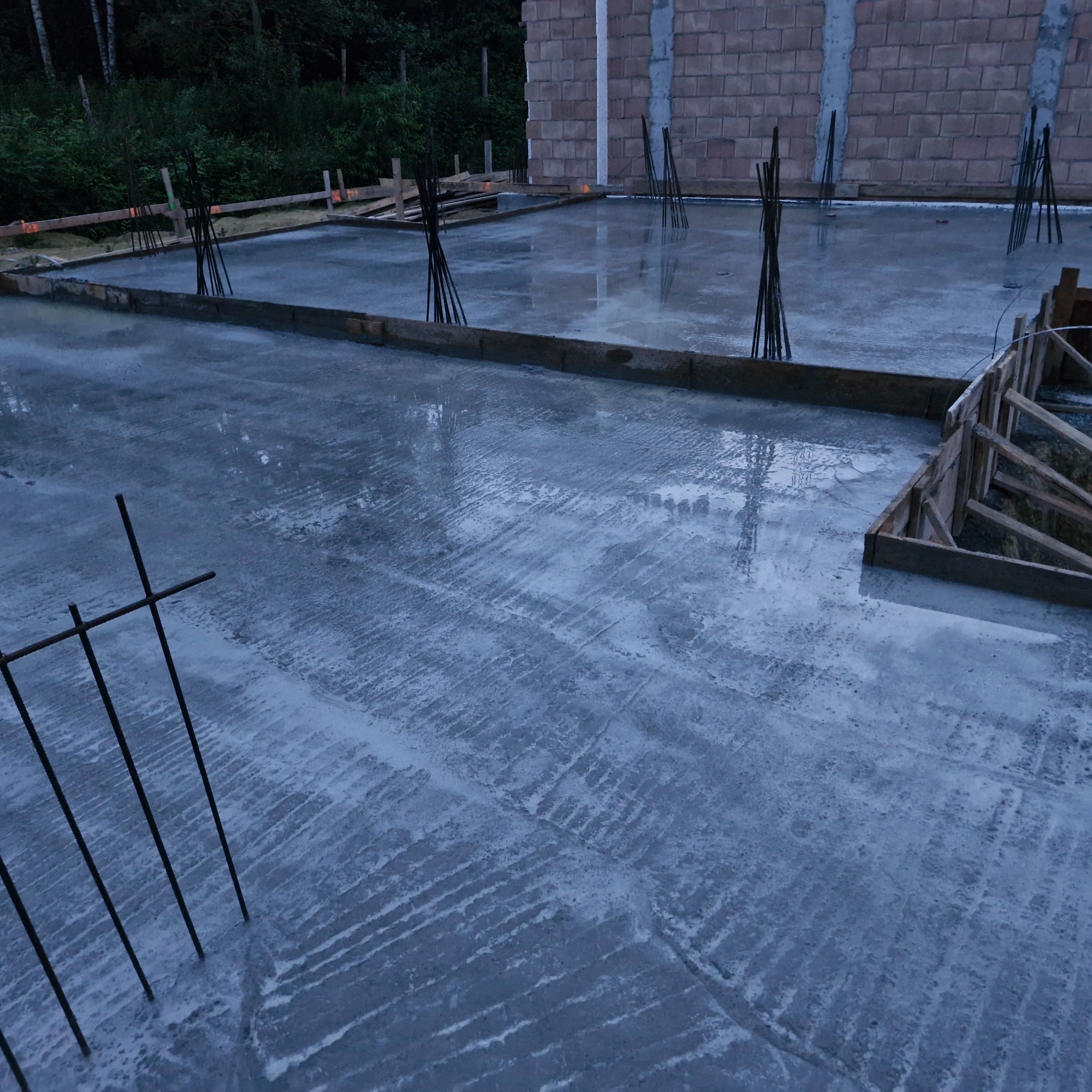 Zalany betonowy fundament z wystającymi prętami zbrojeniowymi, otoczony drewnianym szalunkiem. W tle ściana z bloczków betonowych i zieleń.