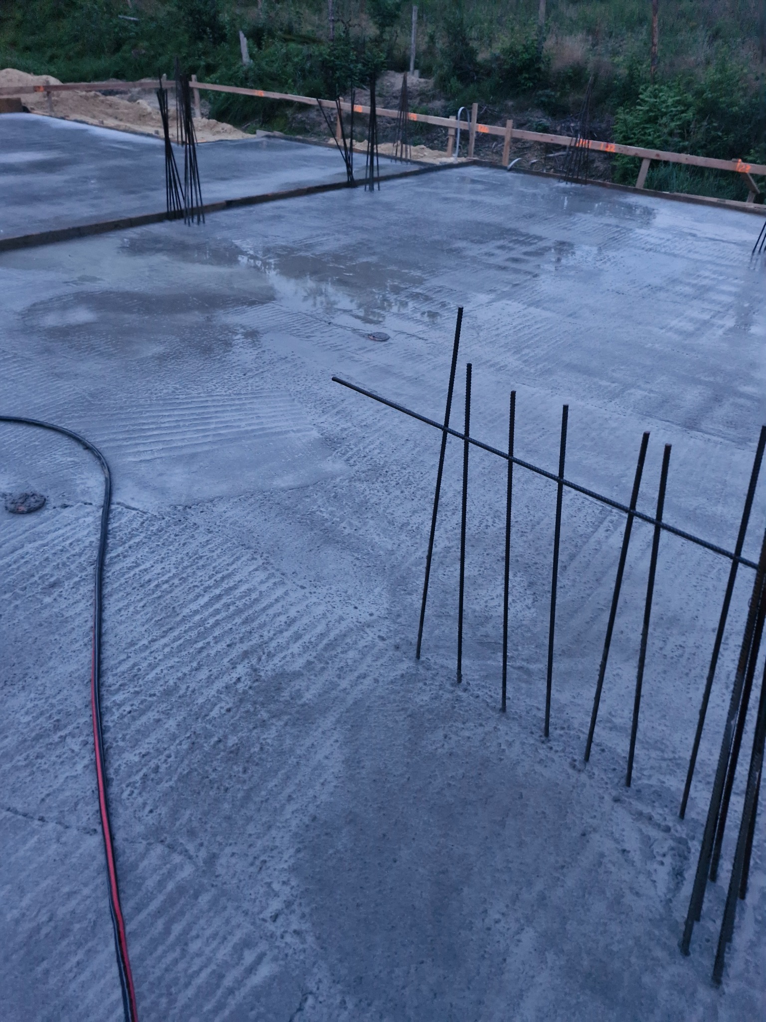 Świeżo wylana płyta fundamentowa z wystającymi prętami zbrojeniowymi, widoczne ślady po szalunku i kabel zasilający po lewej stronie kadru.