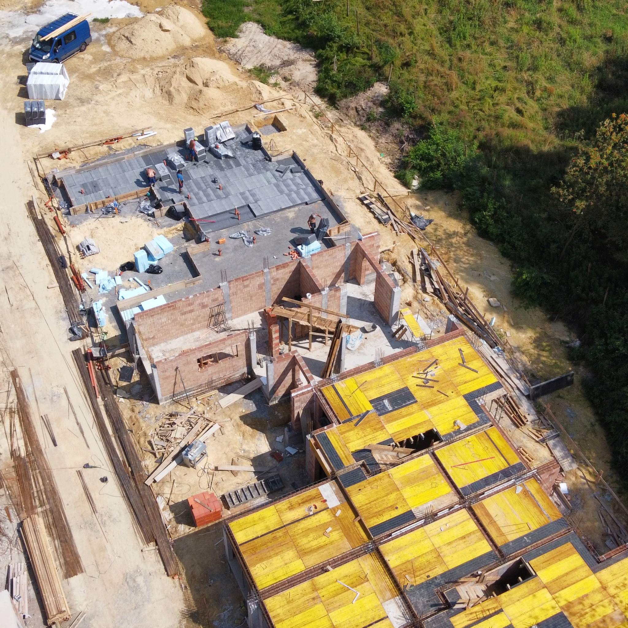 Widok z góry na plac budowy z fundamentami, ścianami z cegły i szalunkami stropowymi. Na terenie budowy widoczny bus i materiały budowlane.