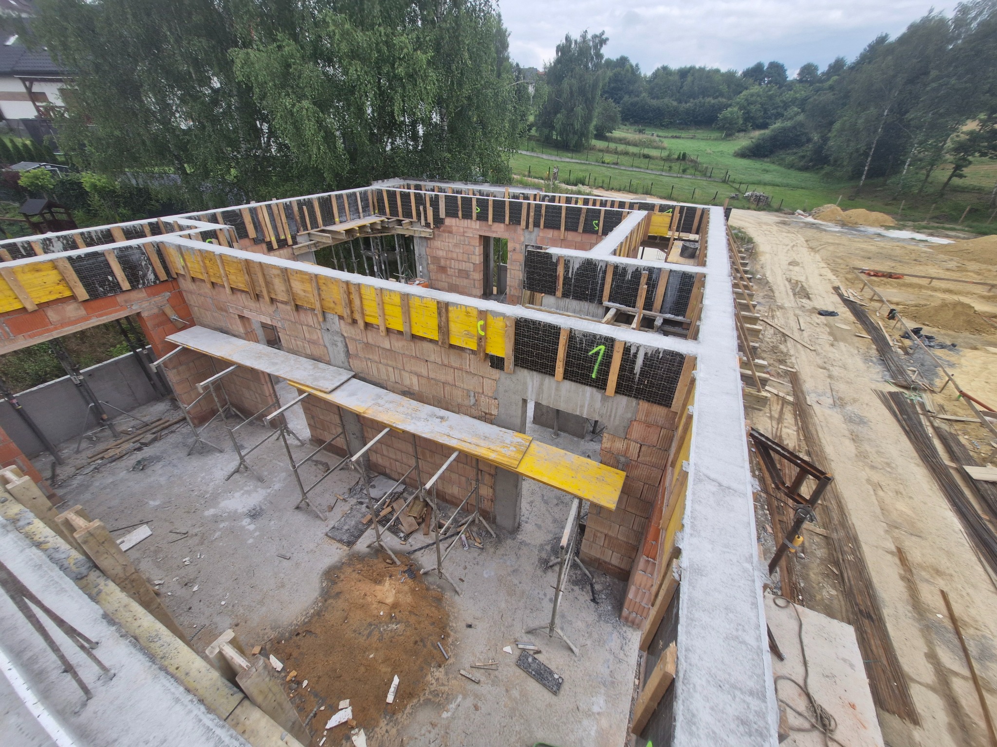 Budowa domu z cegły: zbrojenie wieńca i ściany, żółte szalunki. Widok z góry na plac budowy z zielonym tłem.