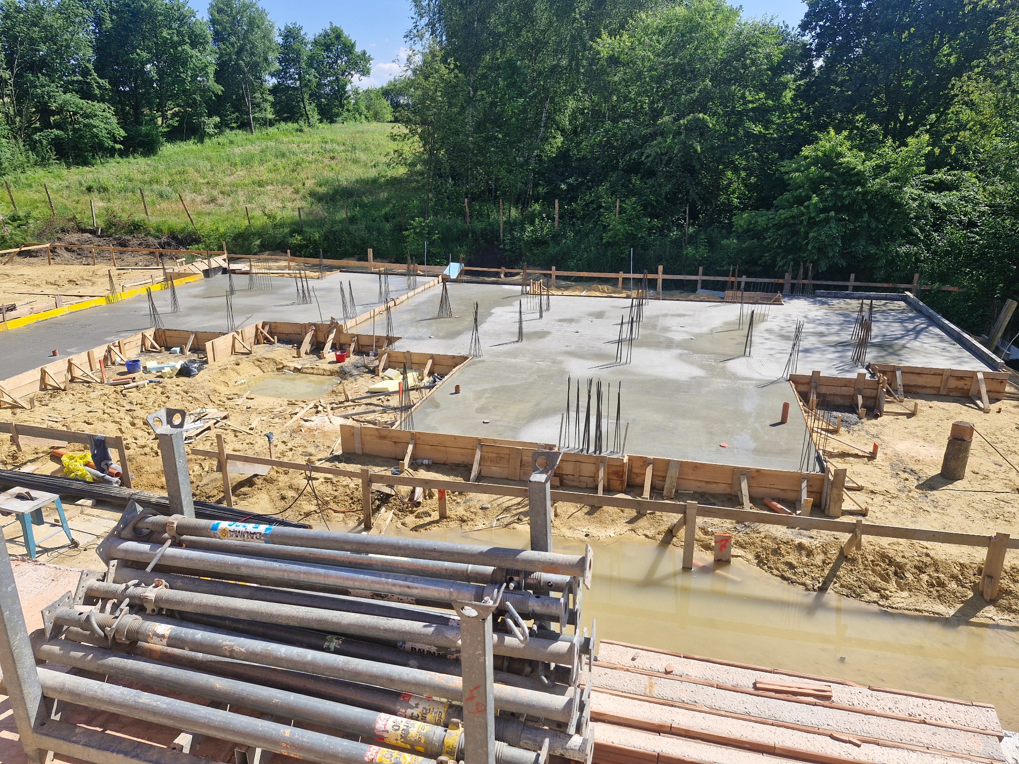 Zalane fundamenty z wystającymi prętami zbrojeniowymi, otoczone drewnianym szalunkiem na placu budowy w słoneczny dzień. W tle zieleń drzew i trawy.