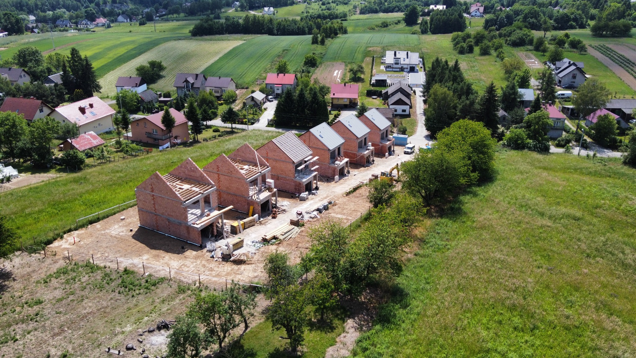 Wysoka perspektywa na szereg domów w trakcie budowy, ceglane ściany, widoczne elementy konstrukcji dachu, otoczenie zieleni i zabudowa wiejska w tle.