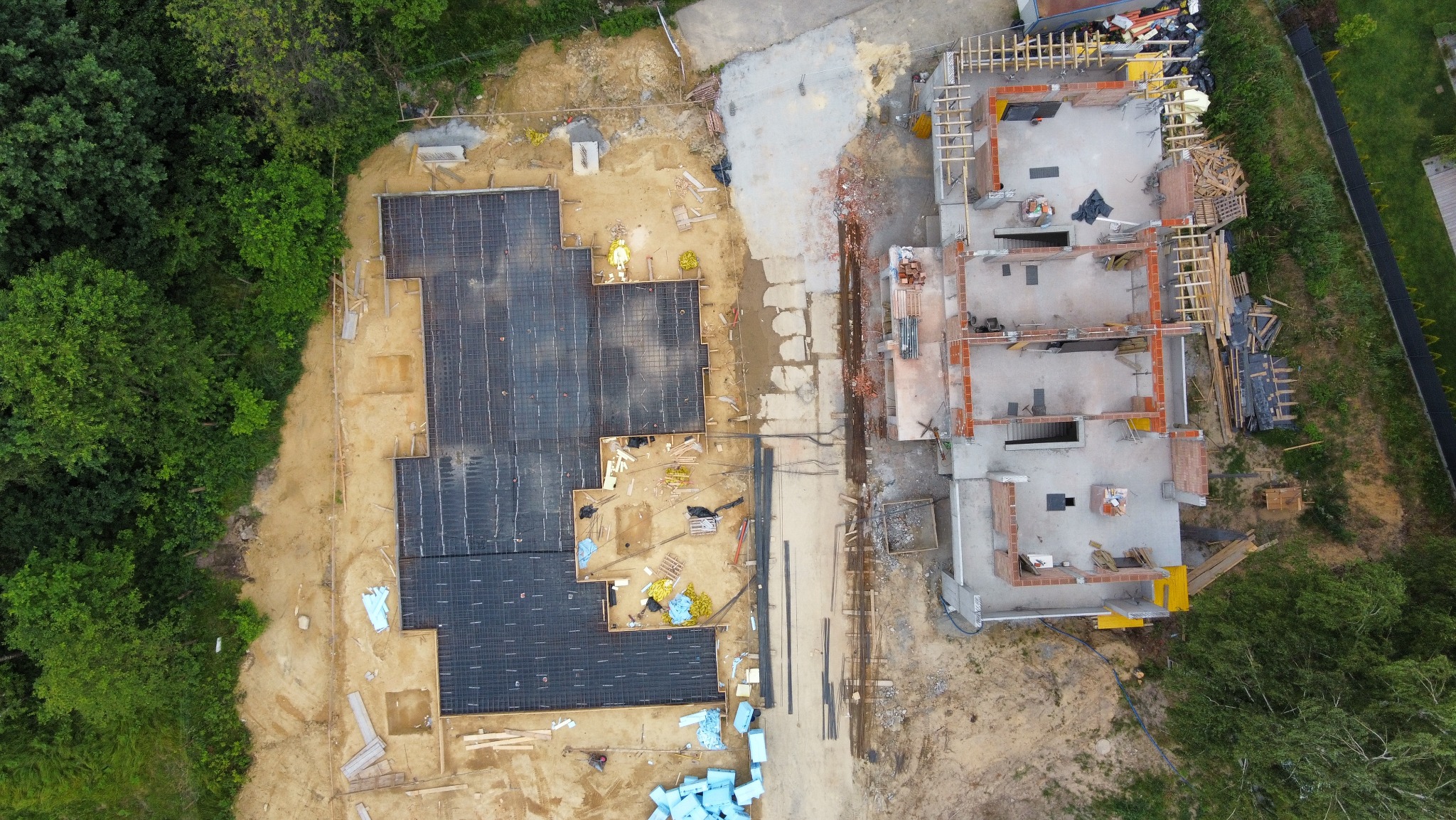 Widok z drona na plac budowy: fundamenty zbrojone na czarno i mury parteru domu z cegły, otoczone zielenią i piaskiem, ukazujące etap prac budowlanych.