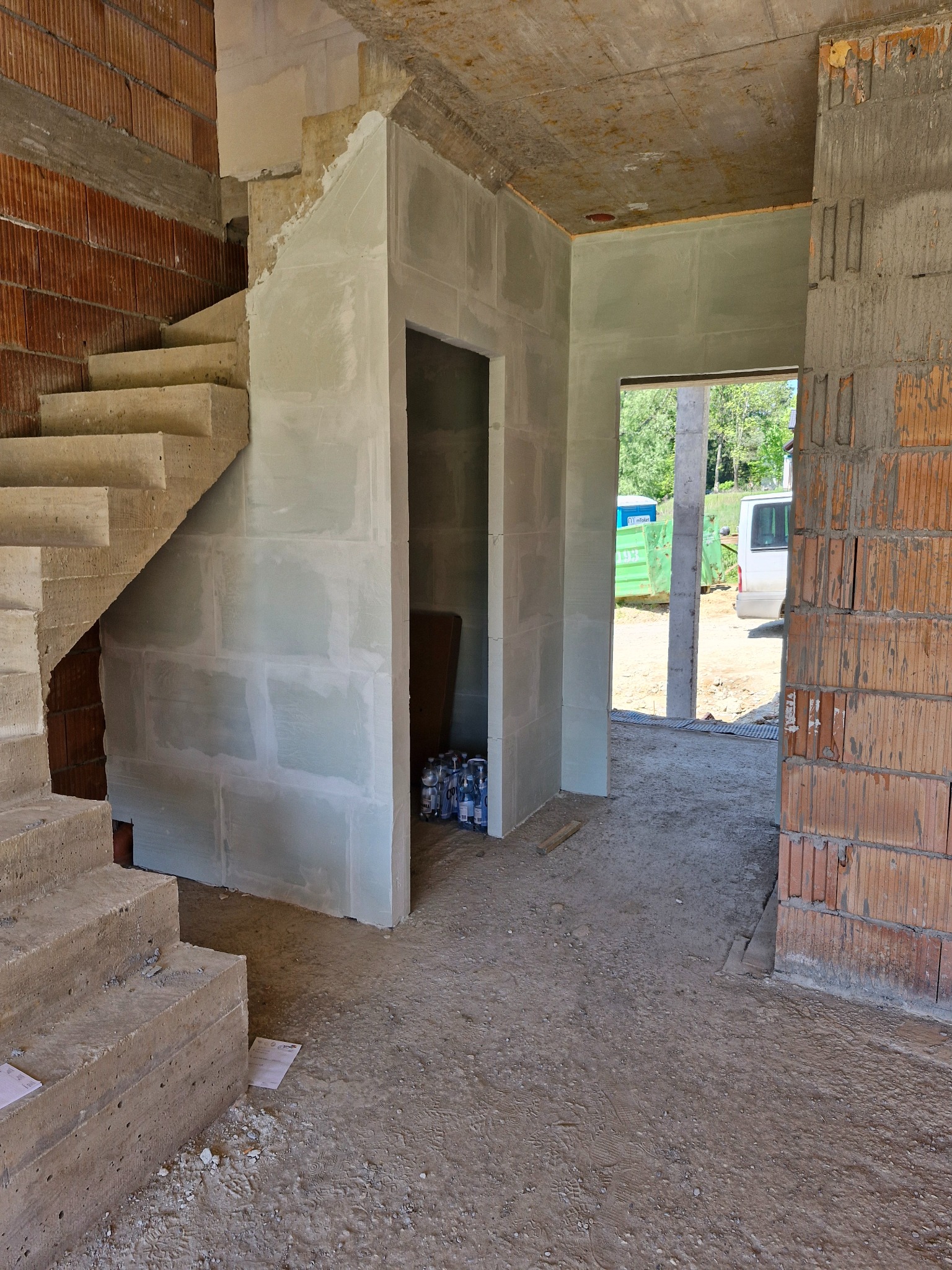 Wnętrze w trakcie budowy: surowe schody betonowe, ściany z pustaków i płyt gipsowych. Widoczny zarys przyszłych pomieszczeń i otwory drzwiowe. Na zewnątrz zieleń i pojazdy.