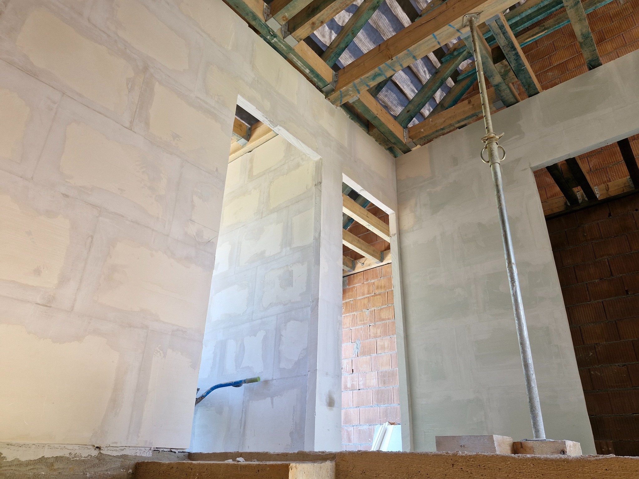 Wnętrze domu w trakcie budowy: surowe ściany z widocznymi warstwami tynku, drewniana konstrukcja dachu i ceglane ściany w tle. Wspornik podtrzymujący strop.