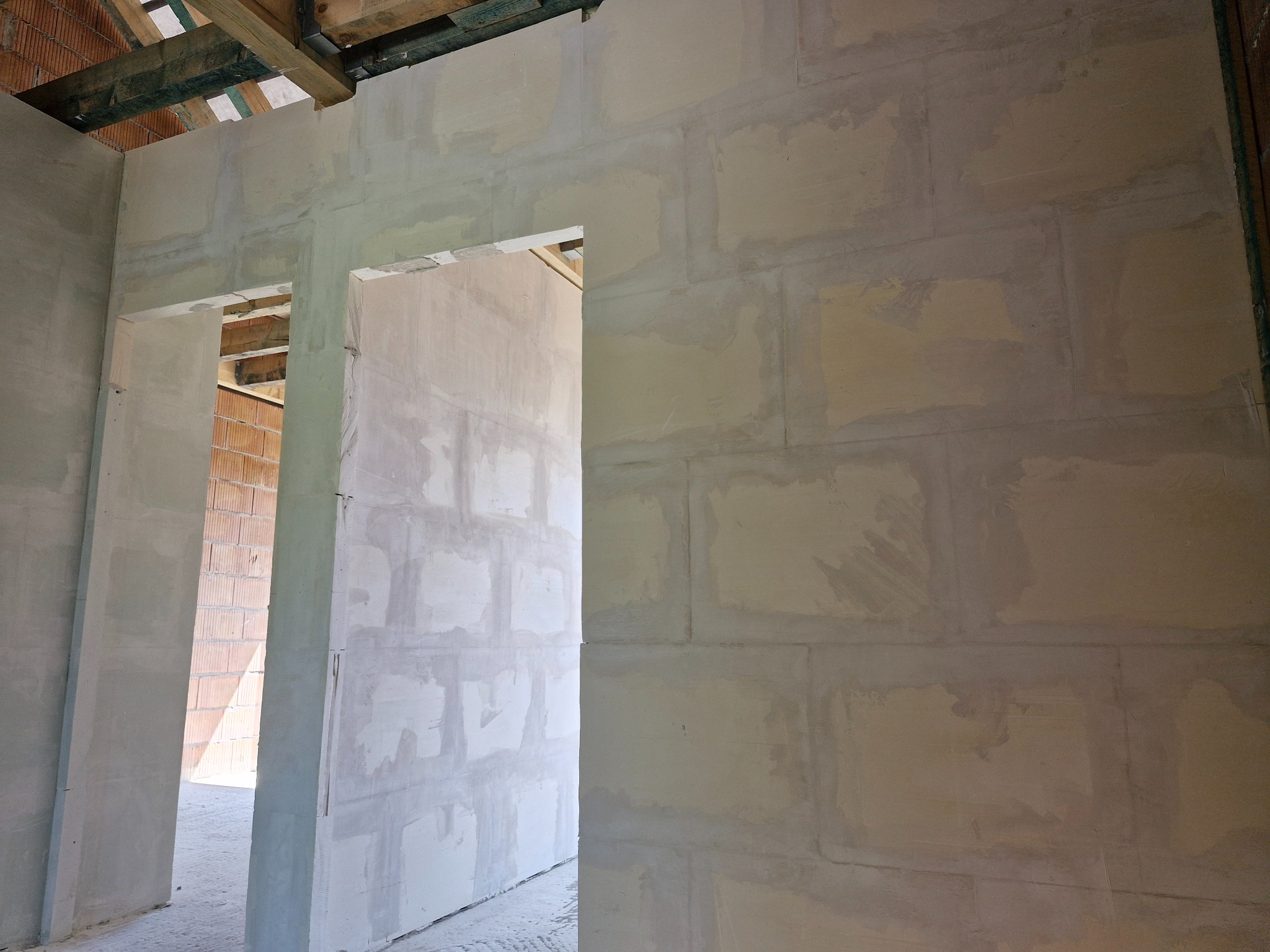 Wnętrze w trakcie budowy: widoczne surowe ściany z betonu komórkowego, częściowo pokryte szpachlą, z otworami na drzwi. Widoczna konstrukcja dachu.