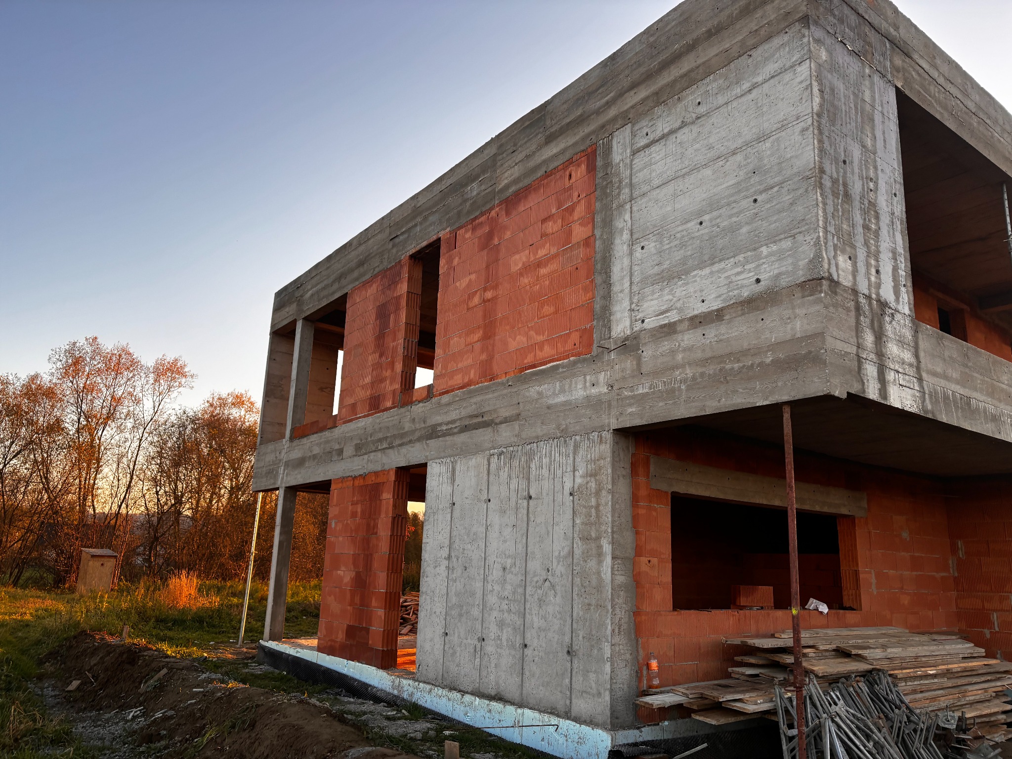 Surowy stan dwukondygnacyjnego domu: betonowe ściany szalunkowe i fragmenty muru z czerwonej cegły. W tle drzewa oświetlone zachodzącym słońcem.