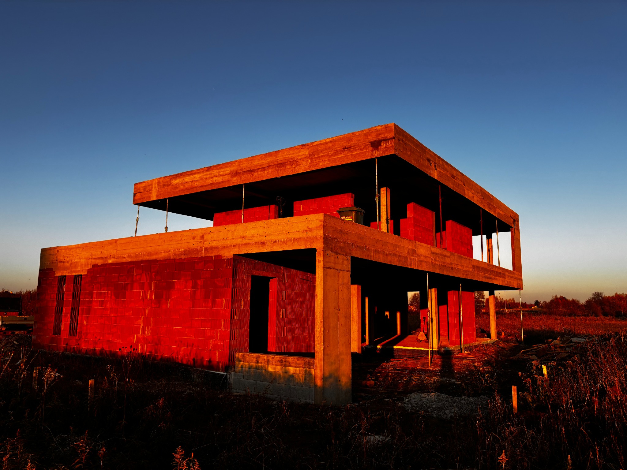 Surowy, dwukondygnacyjny dom z czerwonej cegły silikatowej w trakcie budowy, oświetlony zachodzącym słońcem. Widoczne drewniane szalunki i stalowe podpory.