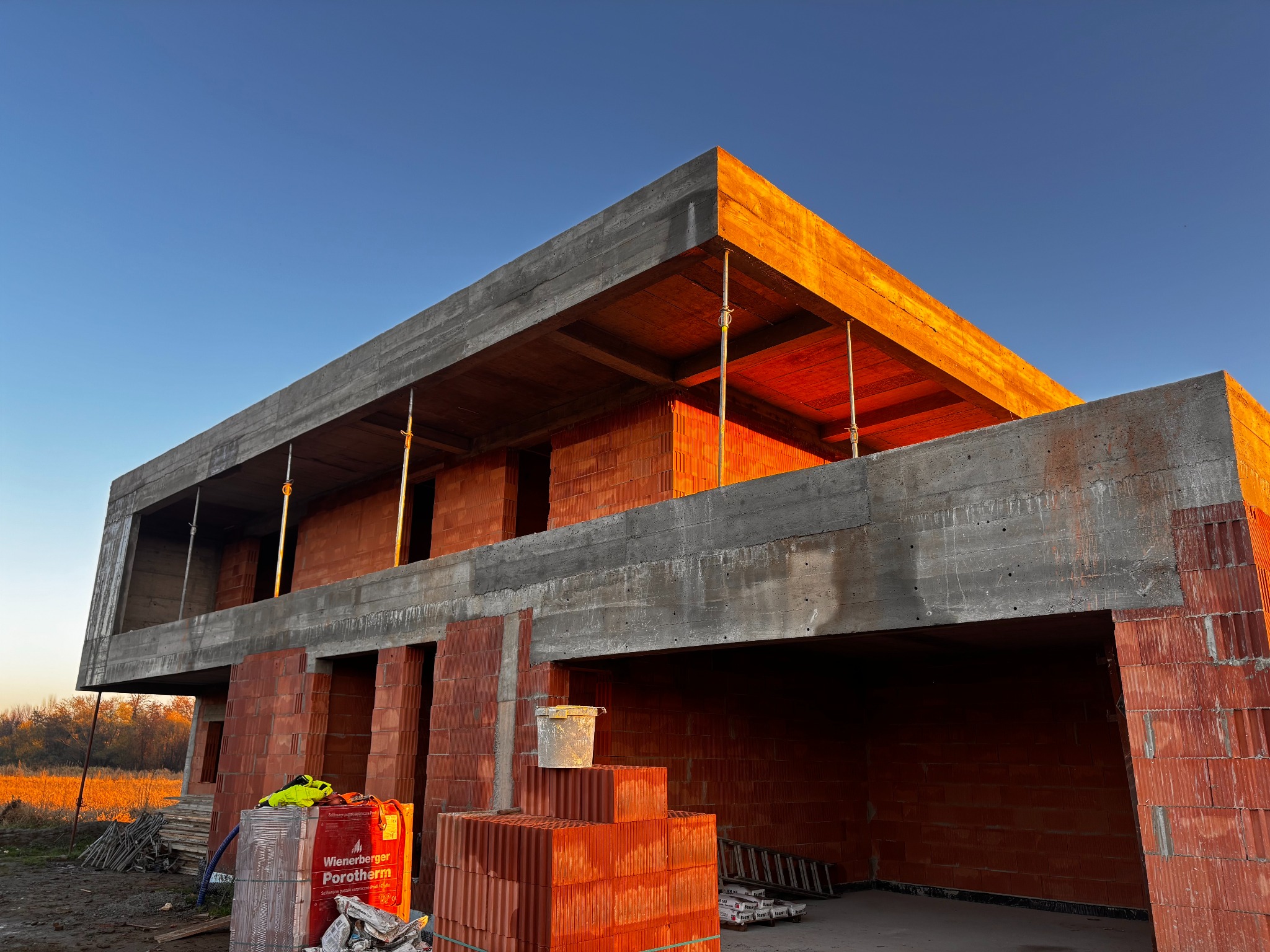 Surowy stan domu z betonu i cegły Porotherm w promieniach zachodzącego słońca. Widoczne podpory stropu i paleta cegieł. Budowa w toku, ciepłe światło.