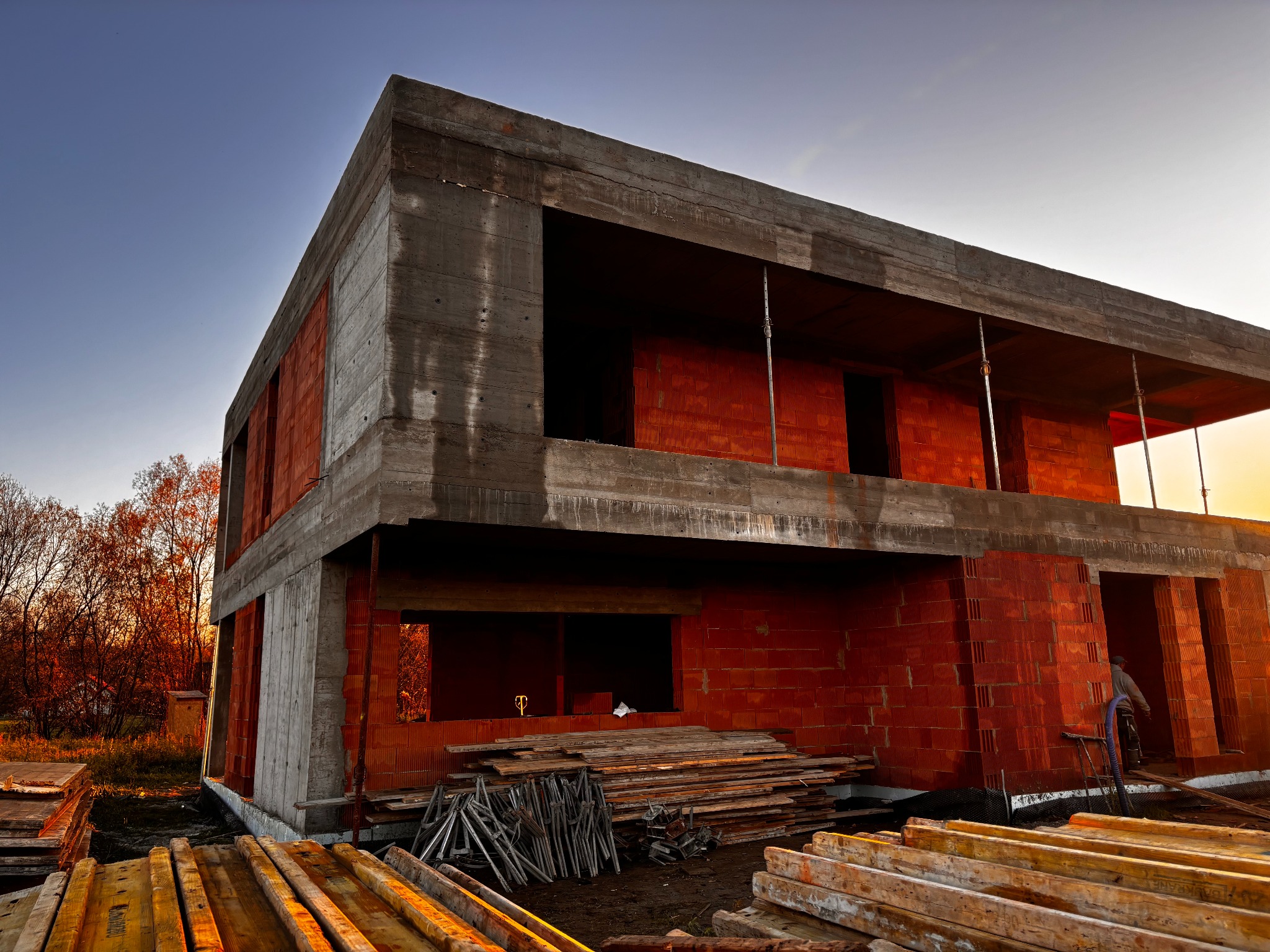 Dwukondygnacyjny budynek w stanie surowym otwartym, z czerwonej cegły i betonu, w otoczeniu drzew i składowanych desek, oświetlony ciepłym światłem zachodzącego słońca.