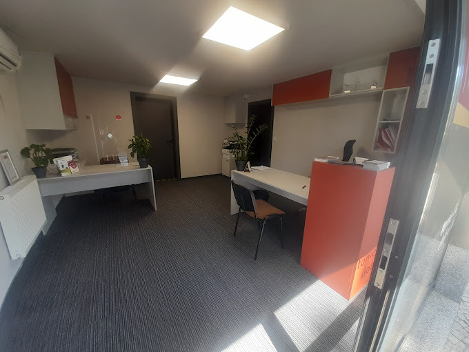 Wnętrze biura z dwoma biurkami, roślinami w doniczkach, szarą wykładziną dywanową i pomarańczowymi akcentami w meblach.