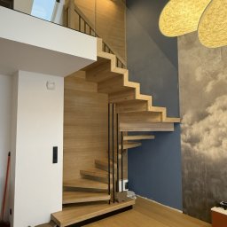 Perfekt-Home - Nowoczesne, drewniane schody wspornikowe z czarną balustradą i szklanym panelem, oświetlone listwą LED, na tle ściany w odcieniach szarości i błękitu.