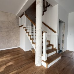 Perfekt-Home - Drewniane schody z białą balustradą i drewnianymi poręczami, wsparte na kolumnie imitującej marmur. Parkiet z naturalnego drewna. Ściana z efektem dekoracyjnym.