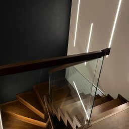 Perfekt-Home - Nowoczesne schody drewniane z balustradą szklaną i drewnianą poręczą, oświetlone ledowymi lampami ściennymi. Widok z góry na ciemne drewno i minimalistyczny design.