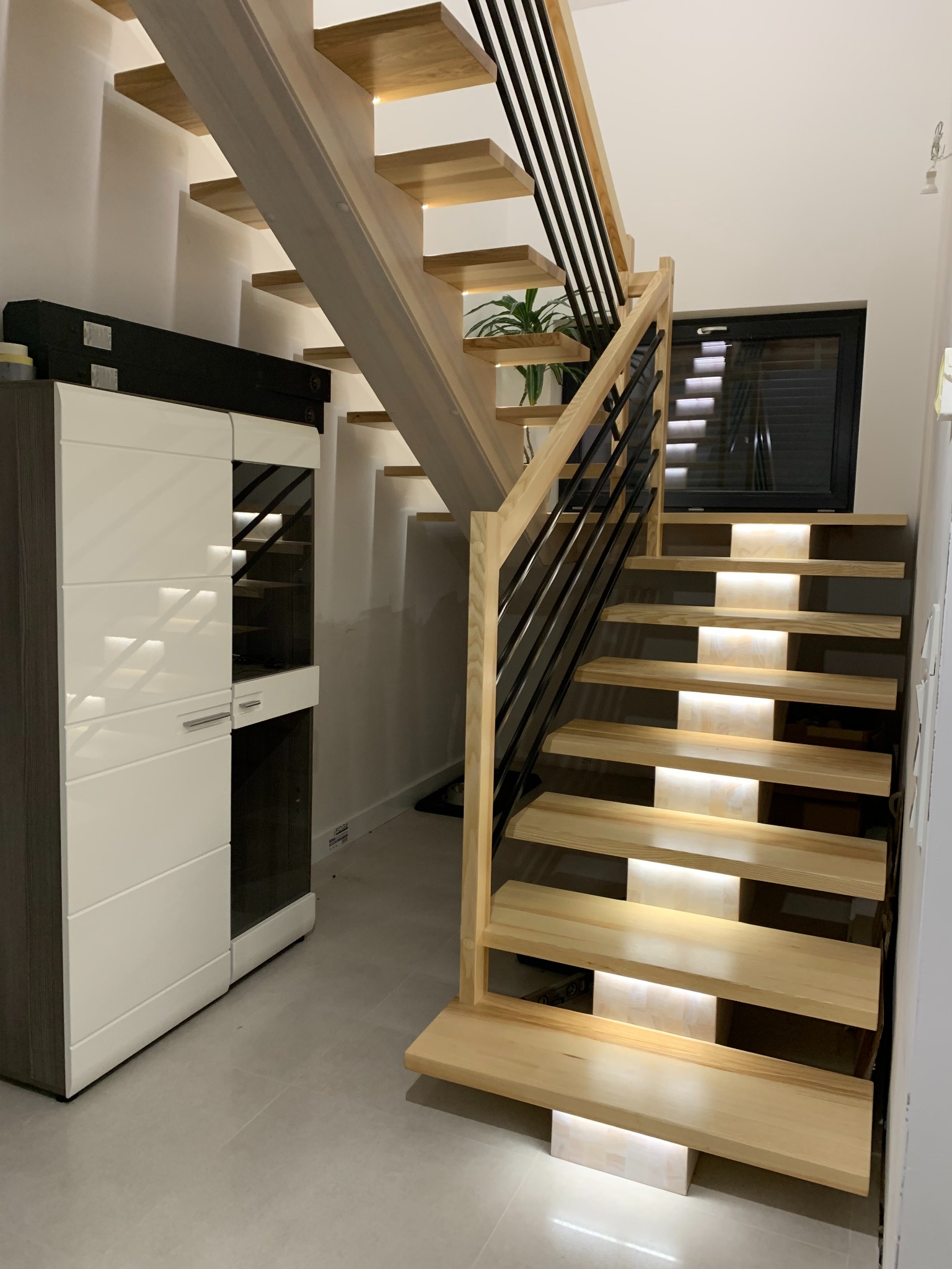 Jasne, drewniane schody z czarną balustradą i subtelnym oświetleniem LED, widziane z dołu. Obok nowoczesna komoda w bieli i szarości. Minimalistyczny design.