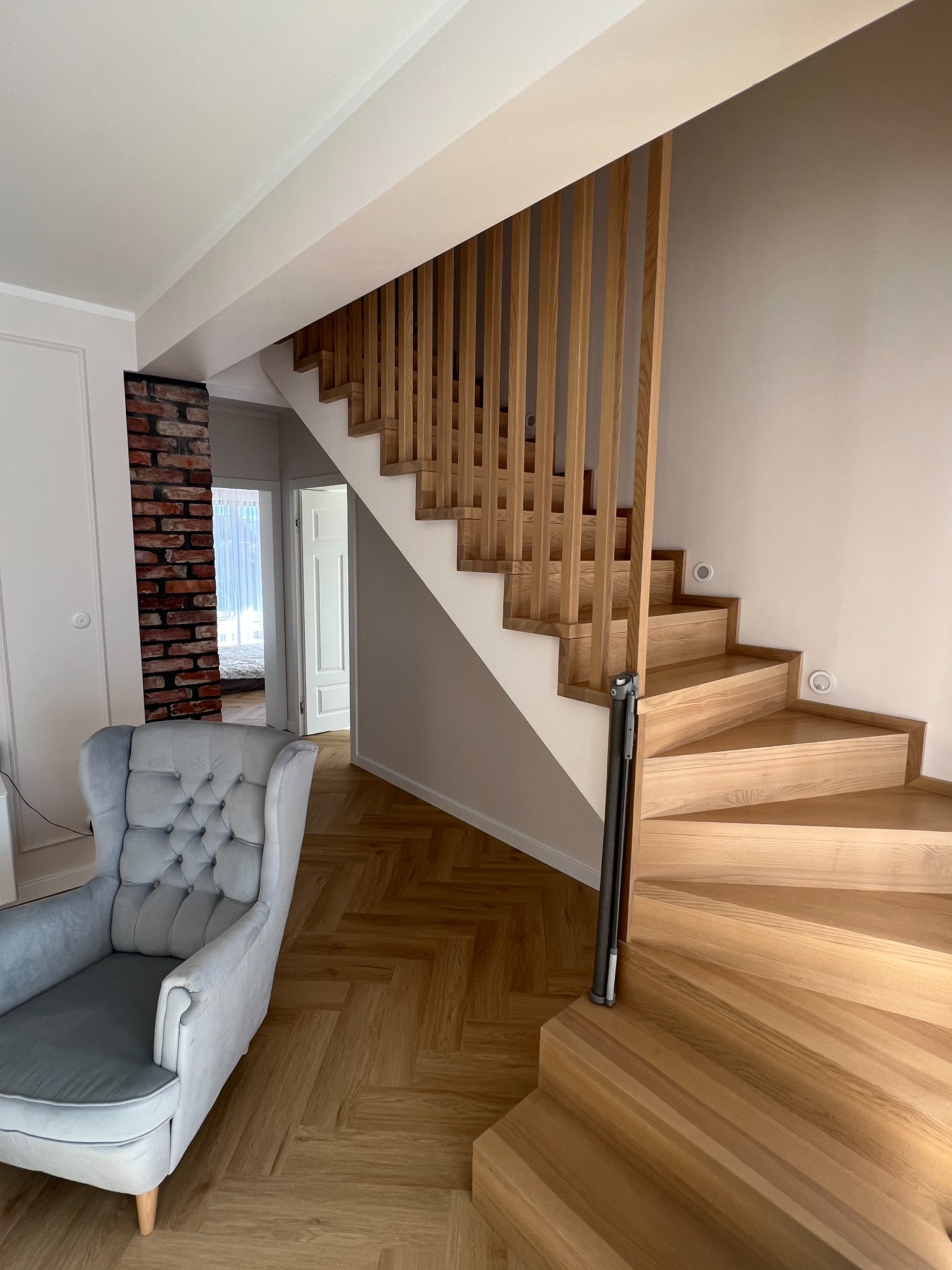 Drewniane schody z ażurową balustradą w jasnym wnętrzu. Widoczny fotel i fragment ściany z cegły. Parkiet ułożony w jodełkę. Nowoczesny design.