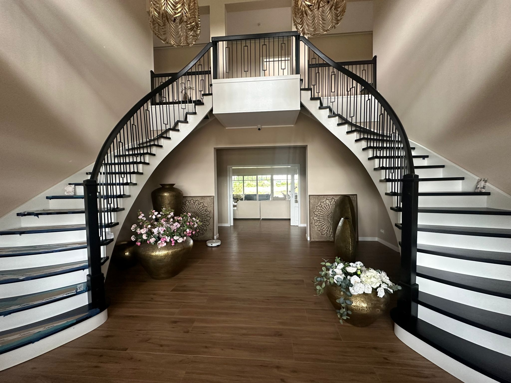 Eleganckie, symetryczne schody z czarną balustradą i białymi stopniami, zdobione kwiatami w złotych wazonach. Parkiet imitujący drewno i dekoracyjne panele na ścianach.