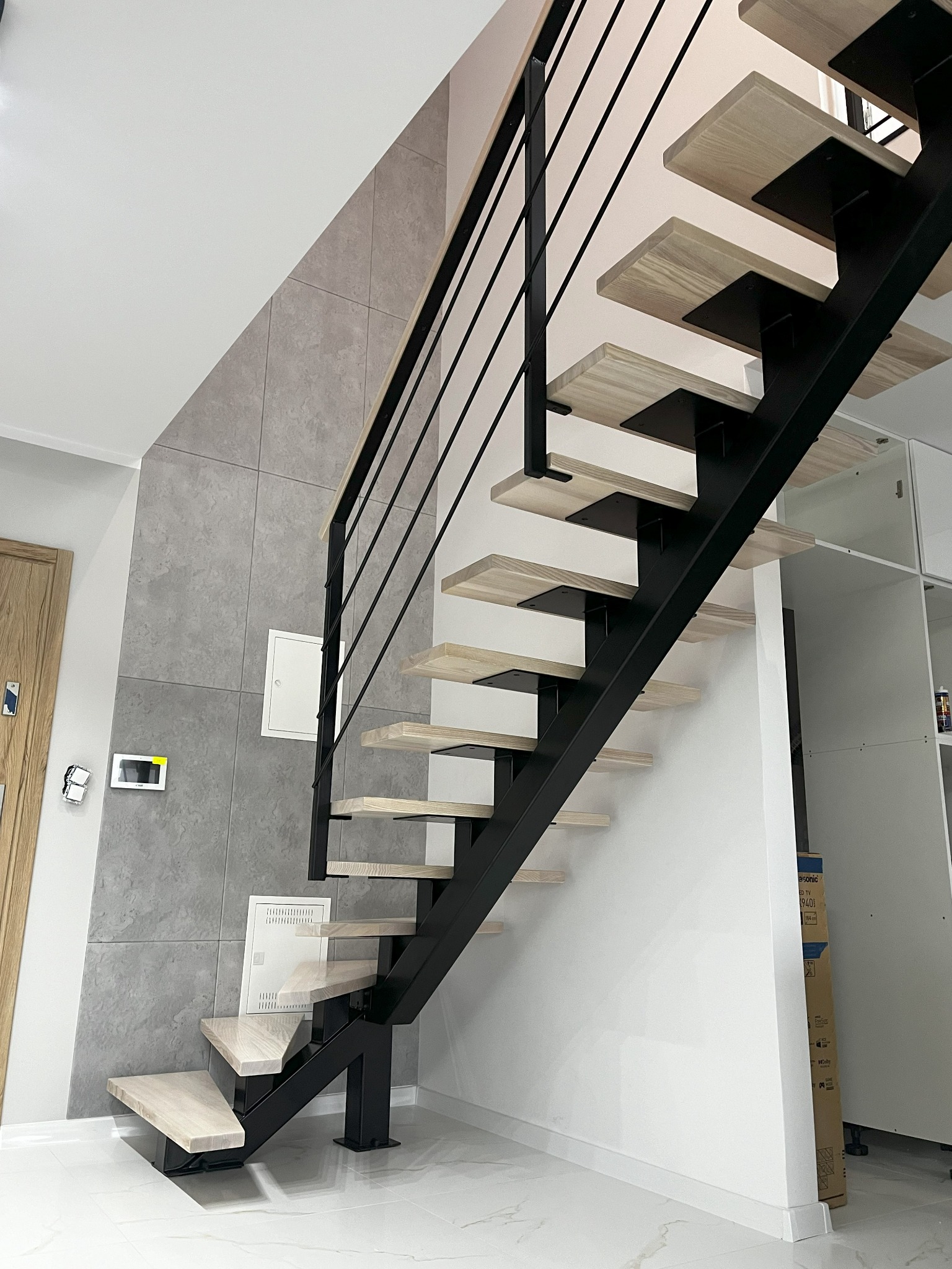 Nowoczesne schody z drewnianymi stopniami i czarną, metalową balustradą. Minimalistyczny design, jasne drewno, szare płytki na ścianie. Widok z dołu, oświetlenie naturalne.