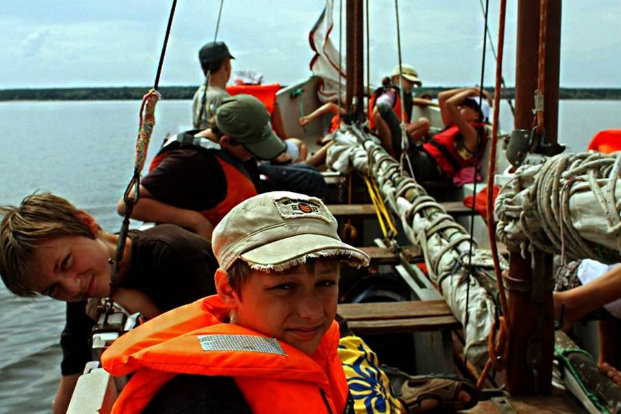 Grupa dzieci w kamizelkach ratunkowych na pokładzie żaglówki podczas rejsu po jeziorze, widok z bliska na chłopca w pomarańczowej kamizelce i czapce z naszywką.