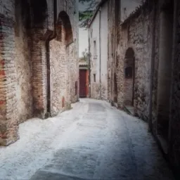 Fototapeta przedstawiająca wąską uliczkę włoskiego miasteczka z kamiennymi budynkami i brukowaną drogą, naklejona na ścianie, z widocznymi panelami podłogowymi u dołu.