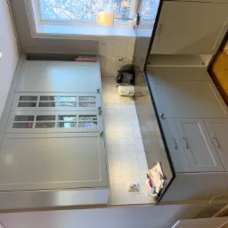 Jasnoszara zabudowa kuchenna z przeszklonymi szafkami górnymi, białe płytki na ścianie, ciemny blat i drewniana podłoga z dywanem w tle.