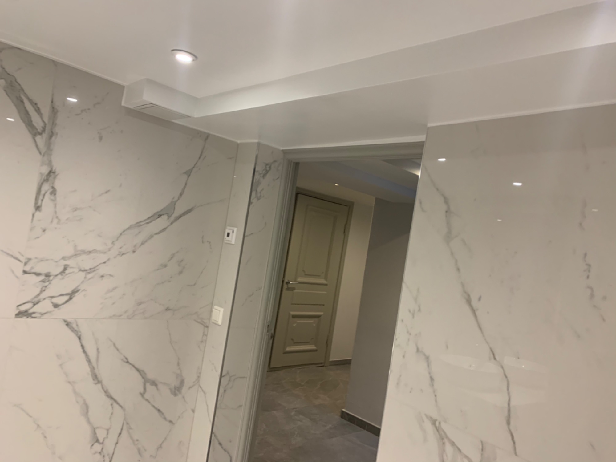 Wykończona łazienka z białymi płytkami imitującymi marmur na ścianach i szarą podłogą, widok na drzwi do innego pomieszczenia.