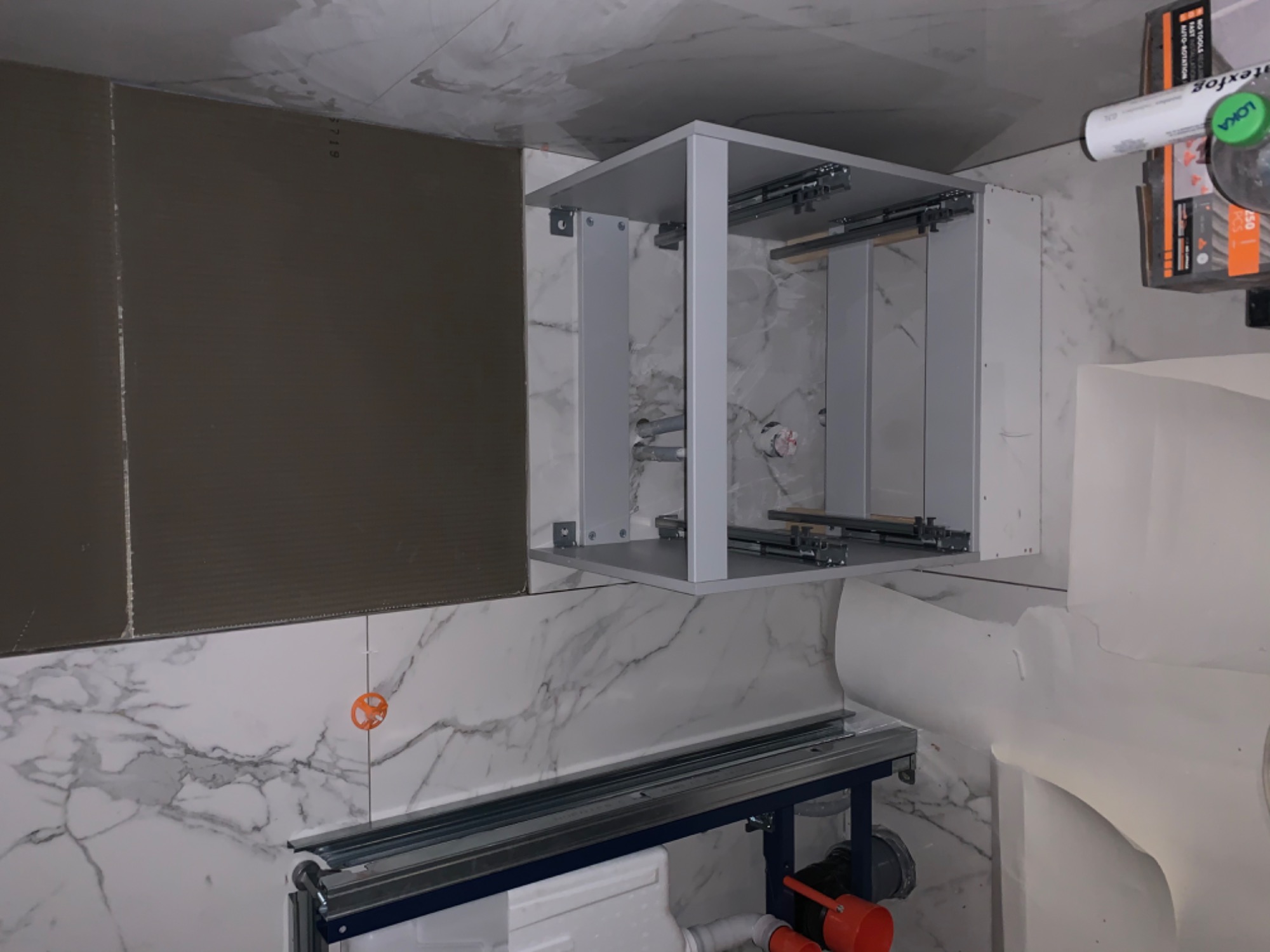 Montaż stelaża podtynkowego WC na tle marmurowych płytek, widoczny element karton gips i szafka z szufladami, w tle opakowanie silikonu.