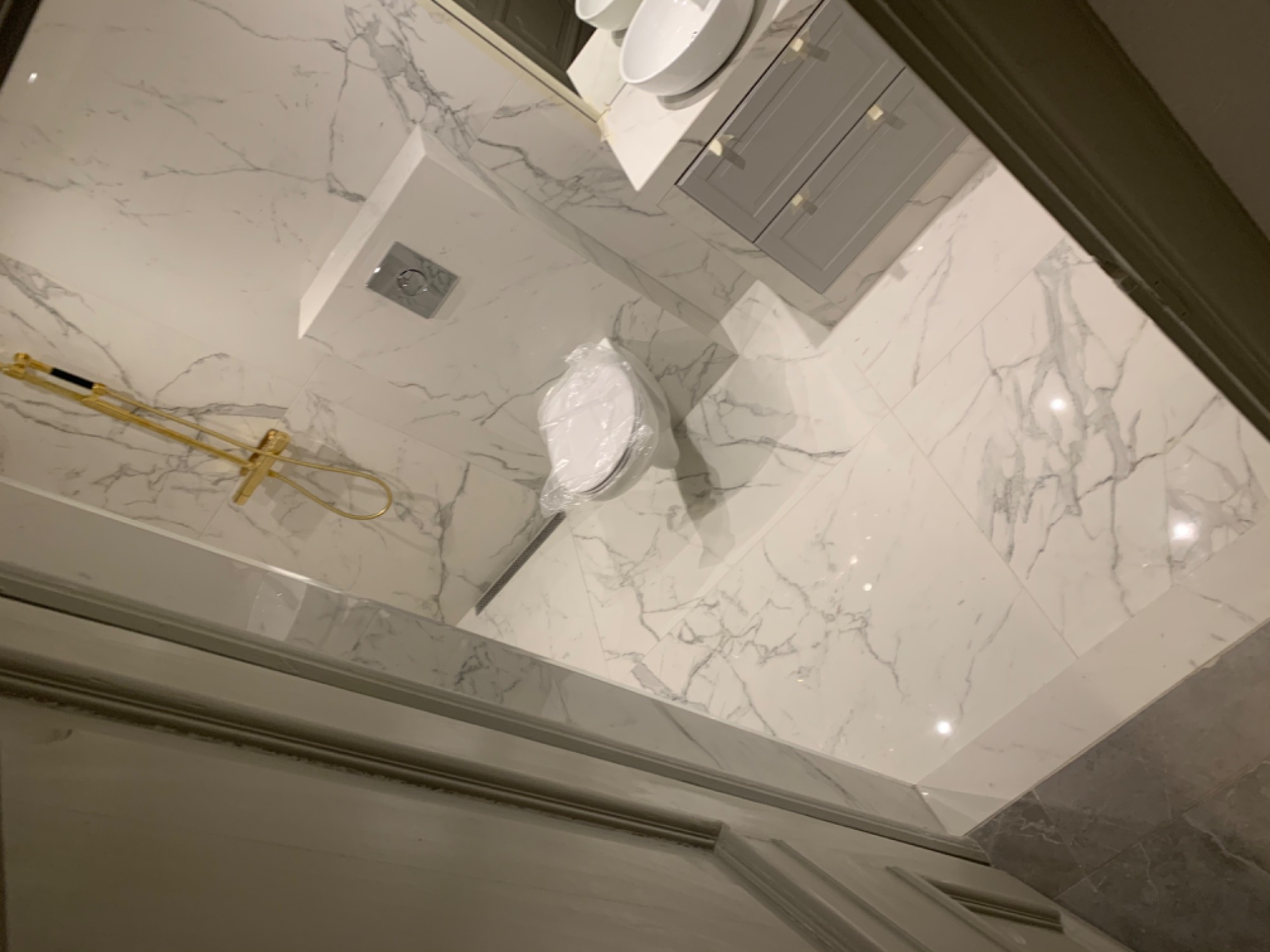 Nowoczesna łazienka z białymi marmurowymi płytkami, złotą armaturą prysznicową i szarą szafką pod umywalką, toaleta jeszcze w folii ochronnej.