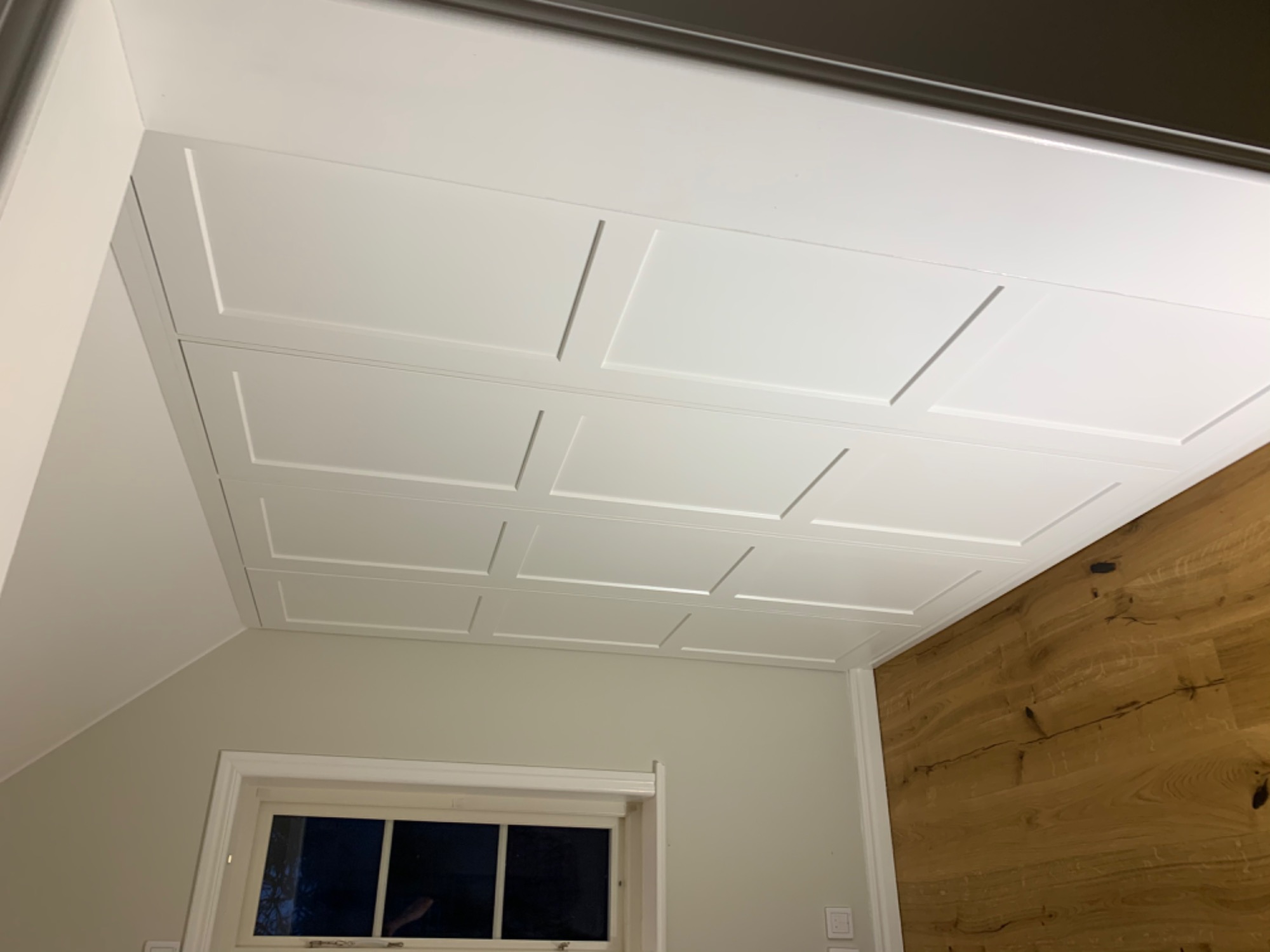 Dekoracyjny, biały sufit kasetonowy z subtelnymi wgłębieniami, widoczny z perspektywy wnętrza, z drewnianą ścianą w tle i oknem po lewej stronie.