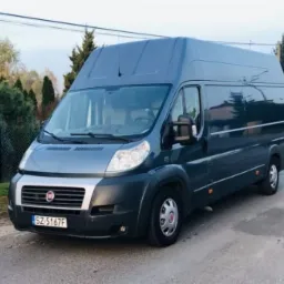 Szary Fiat Ducato z wysokim dachem, zaparkowany na asfaltowej drodze w otoczeniu drzew i krzewów, z widoczną tablicą rejestracyjną SZ 5167F.