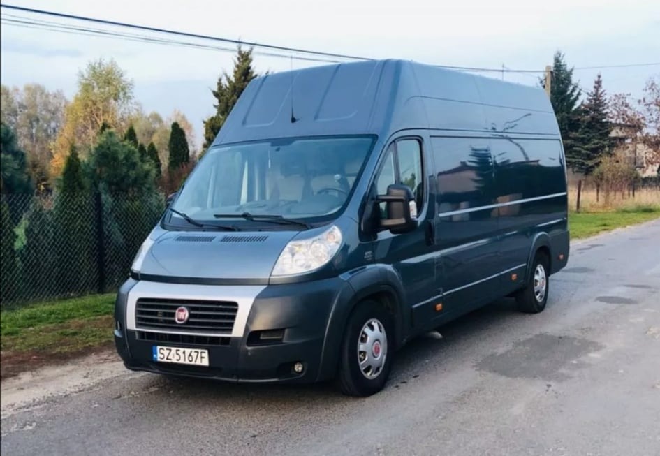 Szary Fiat Ducato z wysokim dachem, zaparkowany na asfaltowej drodze w otoczeniu drzew i krzewów, z widoczną tablicą rejestracyjną SZ 5167F.