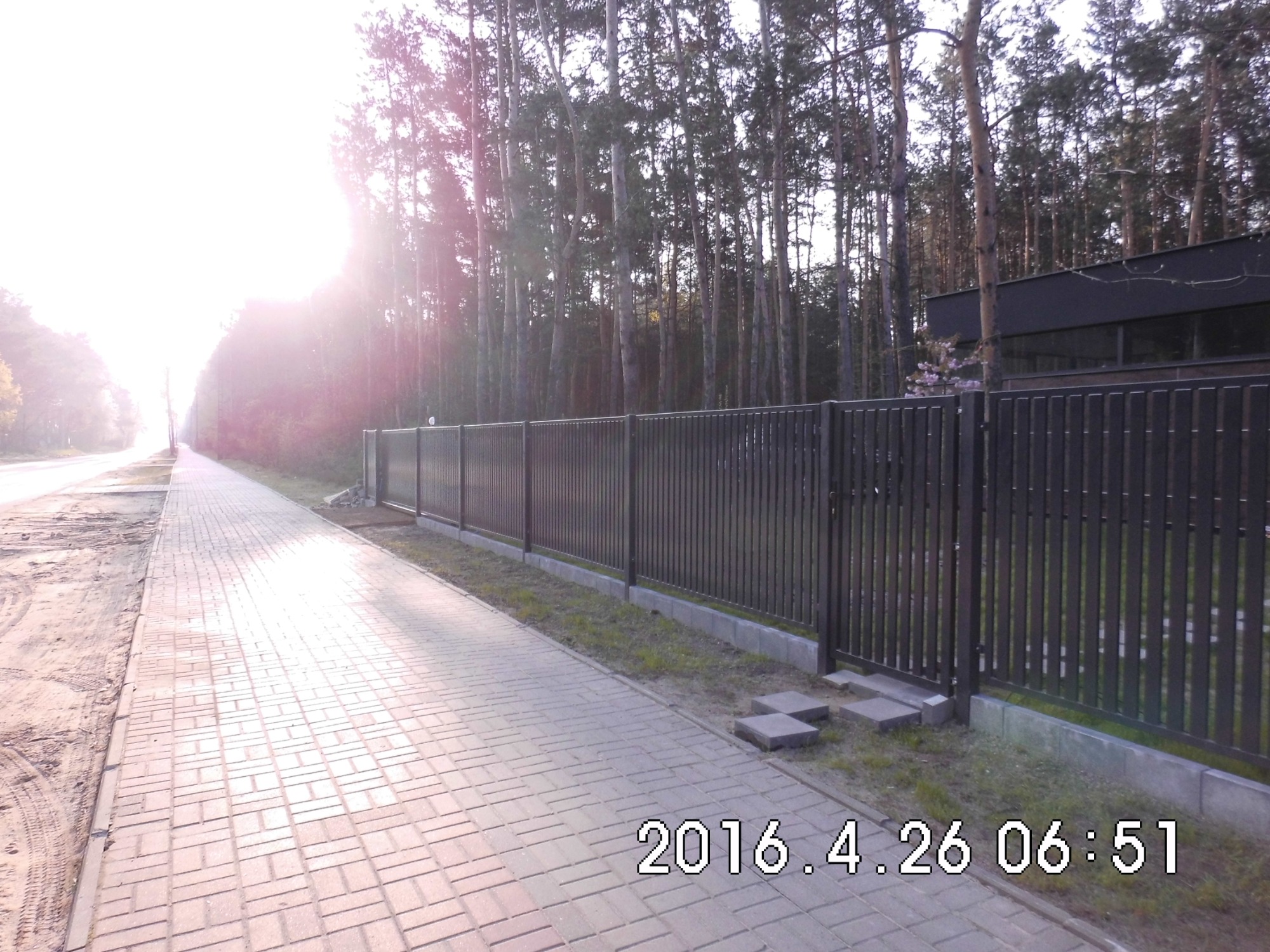 Długa, pionowa, metalowa barierka w kolorze antracytowym, oddzielająca chodnik z kostki brukowej od terenu z drzewami iglastymi i nowoczesnym budynkiem w tle, oświetlona przez wschodzące słońce.