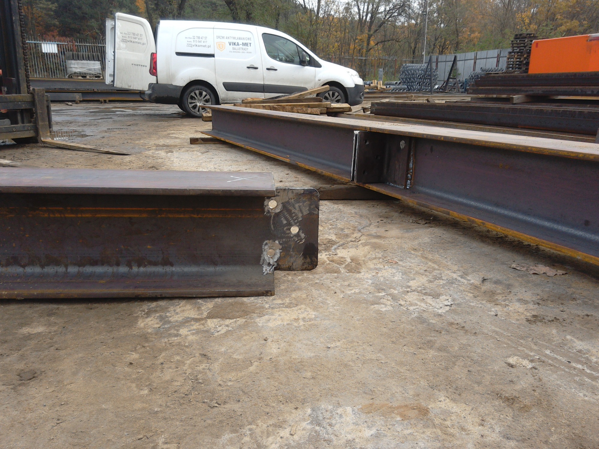 Przygotowane do montażu, spawane stalowe belki dwuteowe z widocznymi spawami, na placu budowy z samochodem dostawczym firmy w tle.