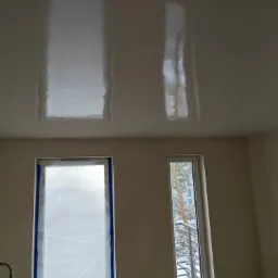 Świeżo położona, lśniąca gładź na suficie w pokoju z oknami zabezpieczonymi folią malarską.