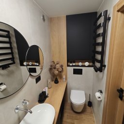 FinishMaster - Nowoczesna łazienka z owalną umywalką nablatową, drewnianym blatem i podłogą w odcieniach drewna, czarnym grzejnikiem drabinkowym oraz okrągłym lustrem z podwójnym odbiciem.