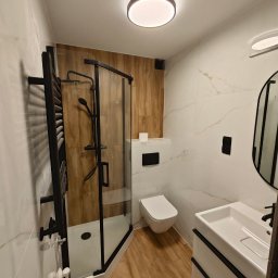 FinishMaster - Nowoczesna łazienka z kabiną prysznicową typu walk-in z czarnymi profilami, umywalką nablatową, podwieszaną toaletą oraz płytkami imitującymi marmur i drewno.