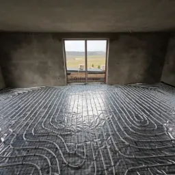 Szare wnętrze w trakcie montażu ogrzewania podłogowego z widocznymi rurami na czarnej siatce, surowe ściany w tle i okno z widokiem na zewnątrz.